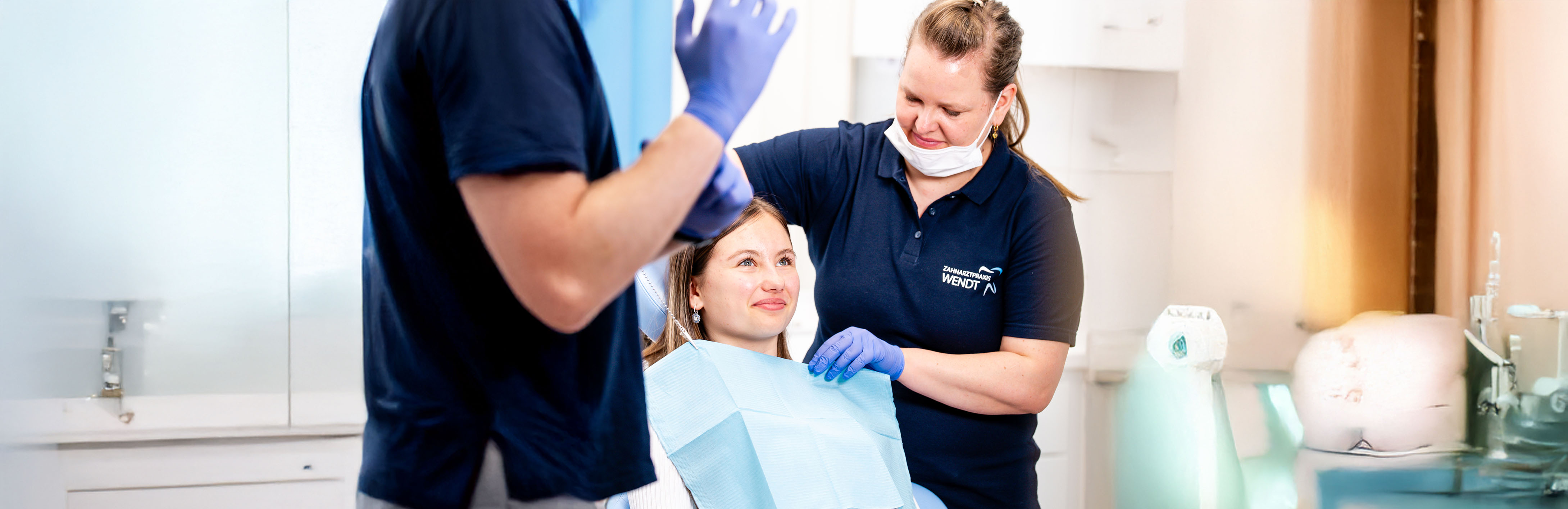 Zahnärztin und Assistent lächeln Patientin an, blau bekleidet, in heller Zahnarztpraxis.