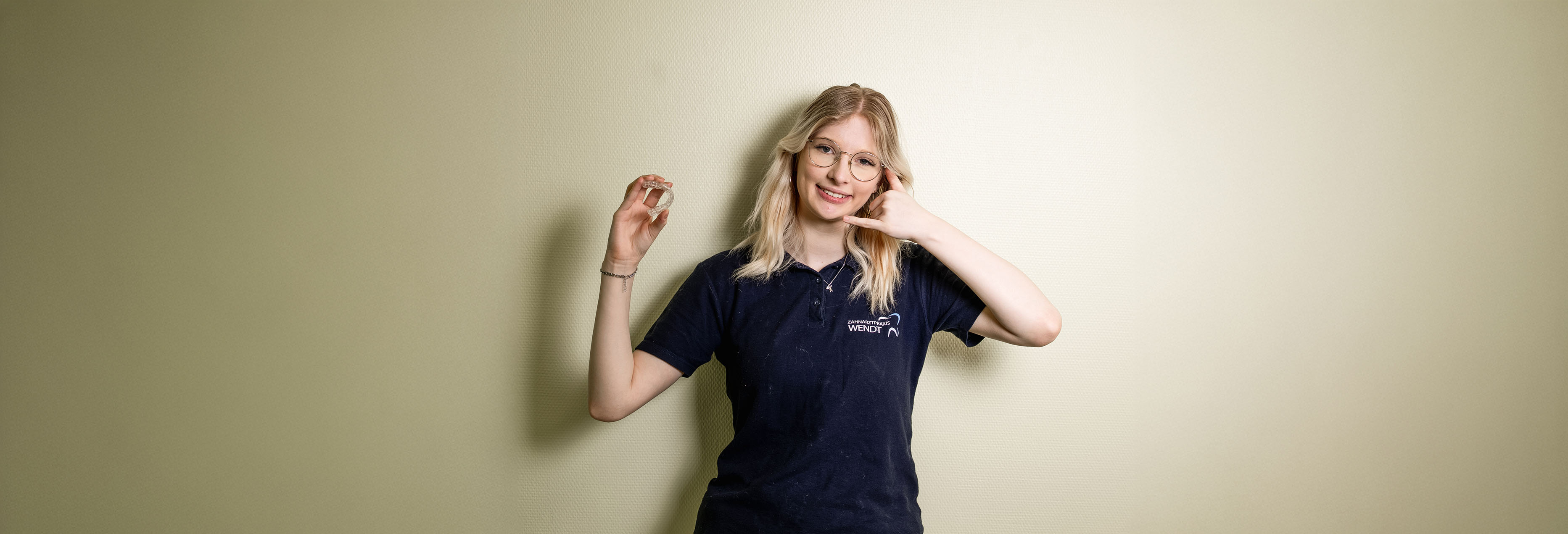 Eine lächelnde Person mit blonden Haaren, die ein Telefonat andeutet, trägt ein dunkelblaues Shirt.