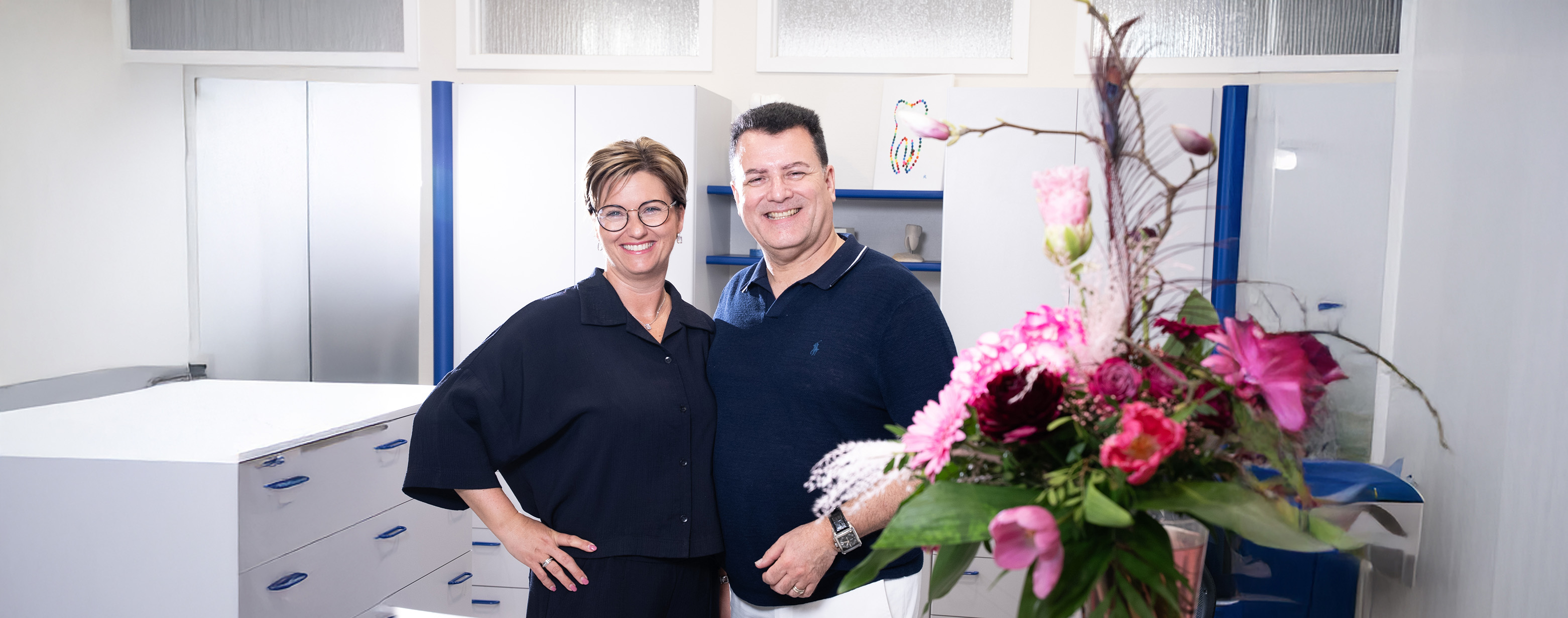 Zwei lächelnde Personen in dunkelblauer Kleidung, in einem hellen Raum mit Blumendekoration.