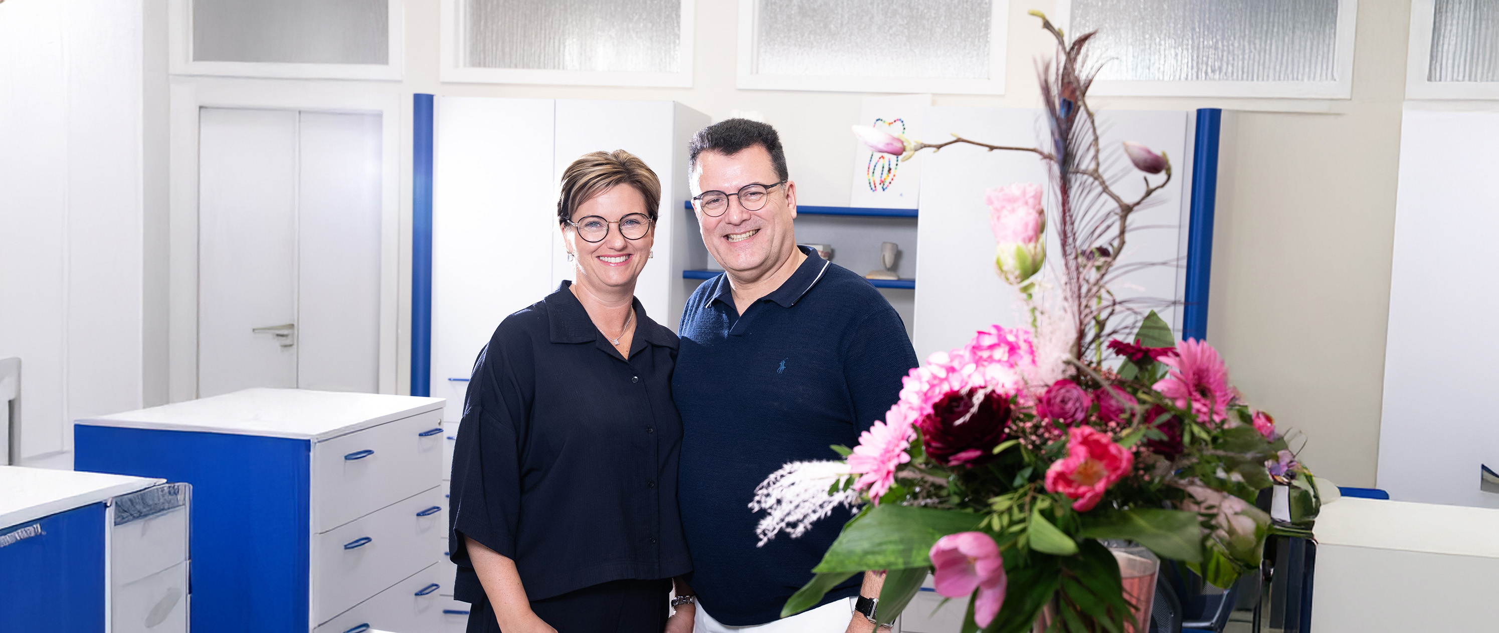 Eine lächelnde Frau und ein Mann in einem Büro mit buntem Blumenstrauß im Vordergrund.