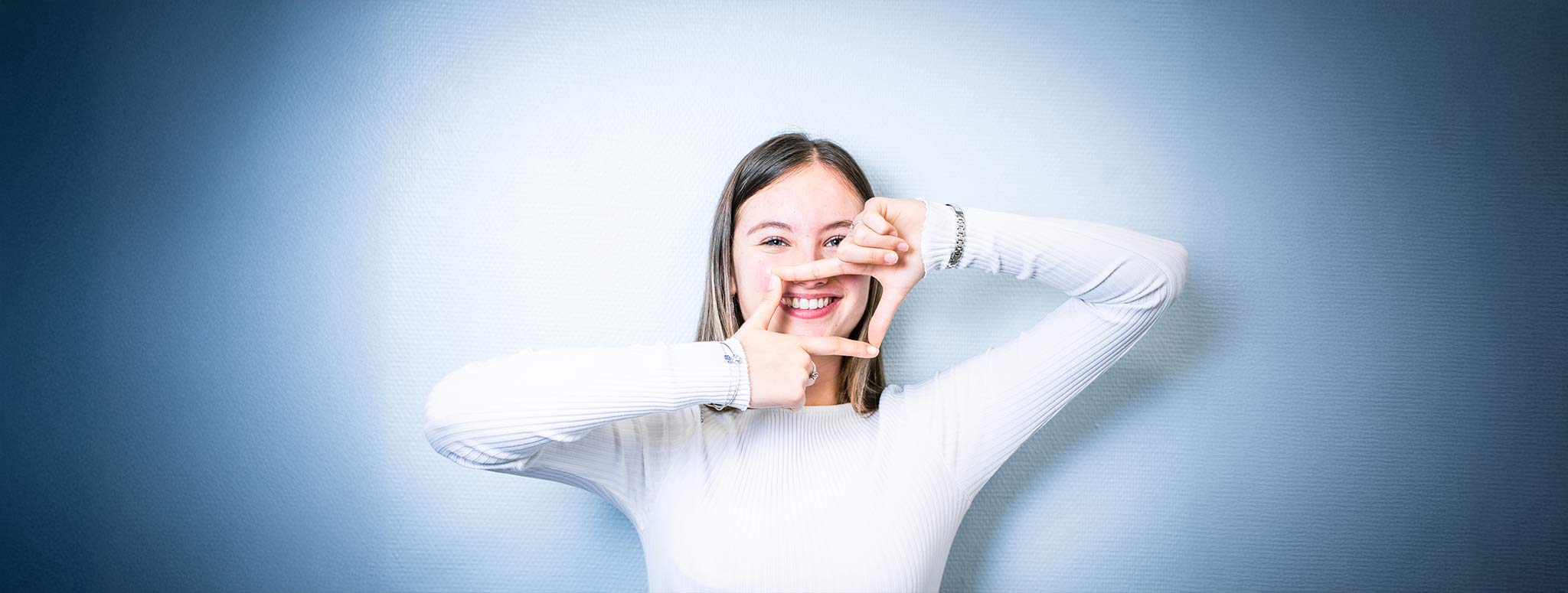 Junge Frau mit hellem Shirt lächelt freundlich und formt mit den Händen ein Rechteck