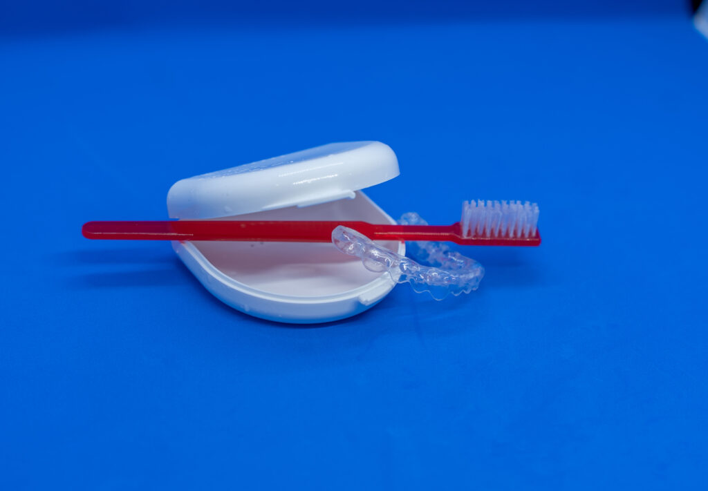 Rote Zahnbürste und transparenter Zahnschutz auf einer weißen Box vor blauem Hintergrund.