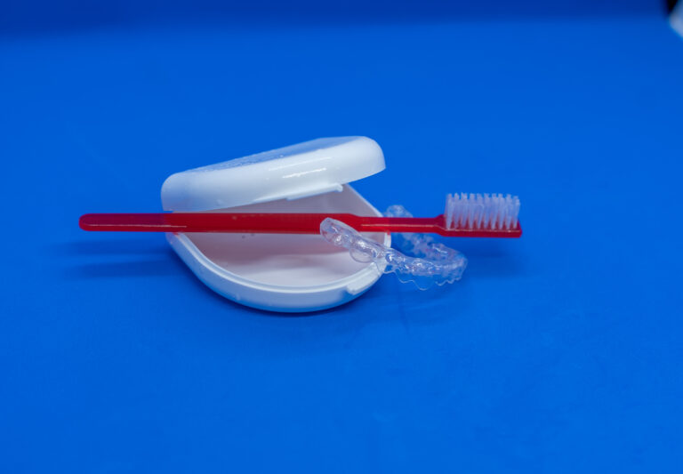 Rote Zahnbürste und transparenter Zahnschutz auf einer weißen Box vor blauem Hintergrund.