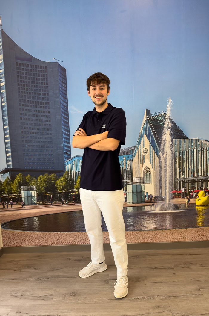 Junger Mann lächelt vor einem Wandbild einer modernen Stadtlandschaft mit Brunnen, trägt dunkles Polo.