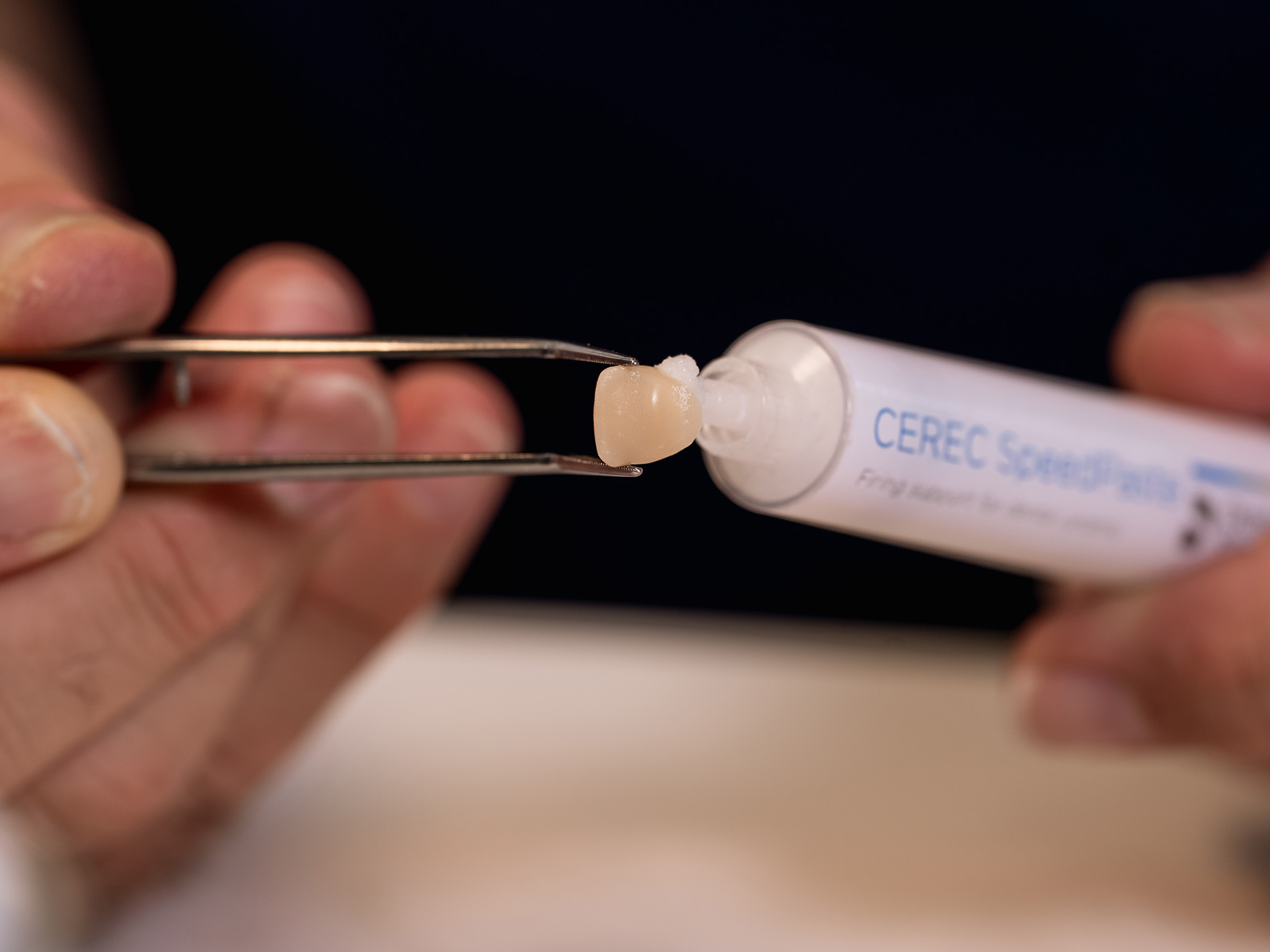 Eine Person hält mit einer Pinzette einen Keramikkern vor einer CEREC SpeedFire-Kapsel.