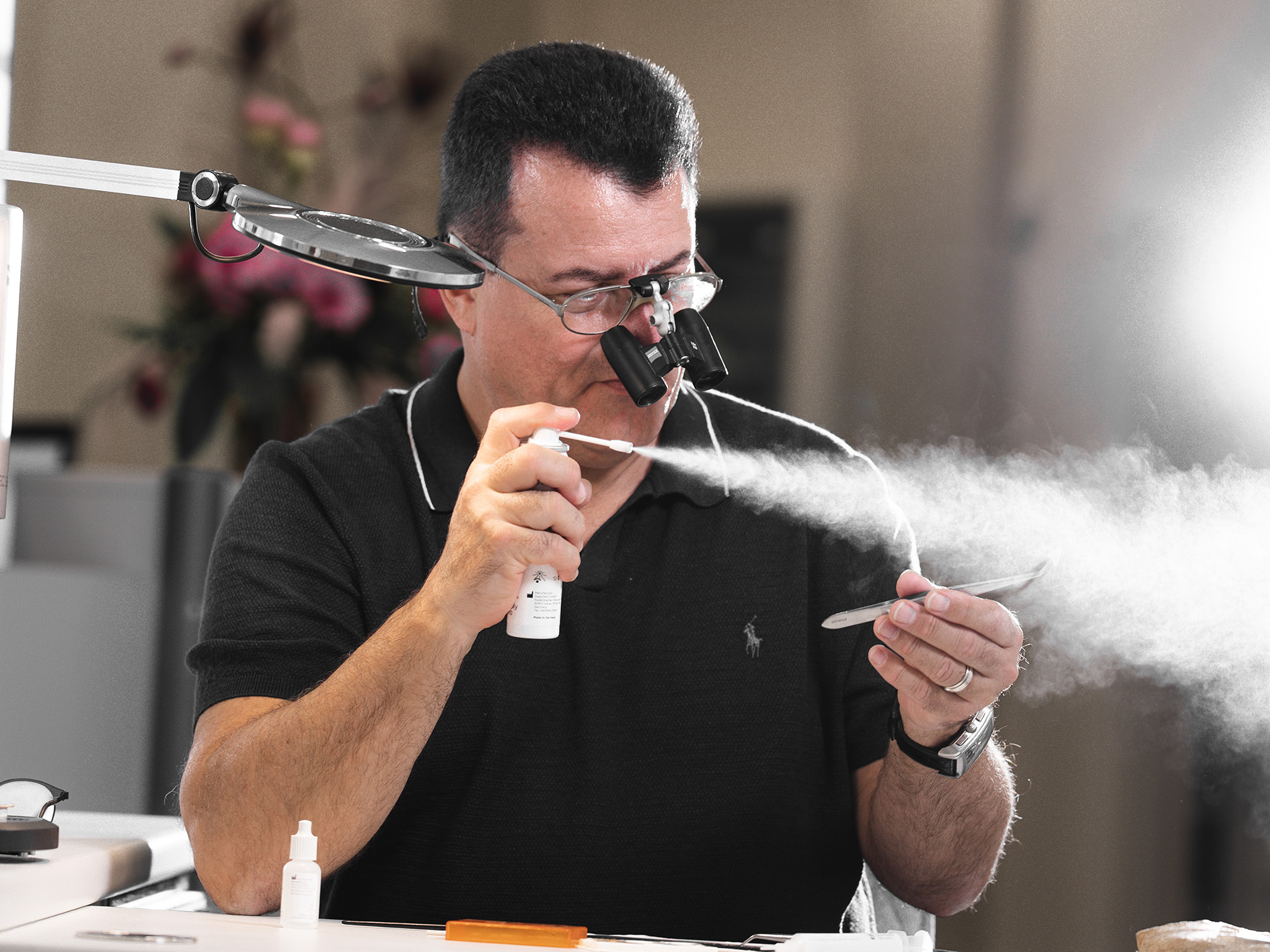 Eine Person konzentriert sich mit Lupenbrille, sprüht Nebel auf Metall-Objekt, trägt dunkles Polo.