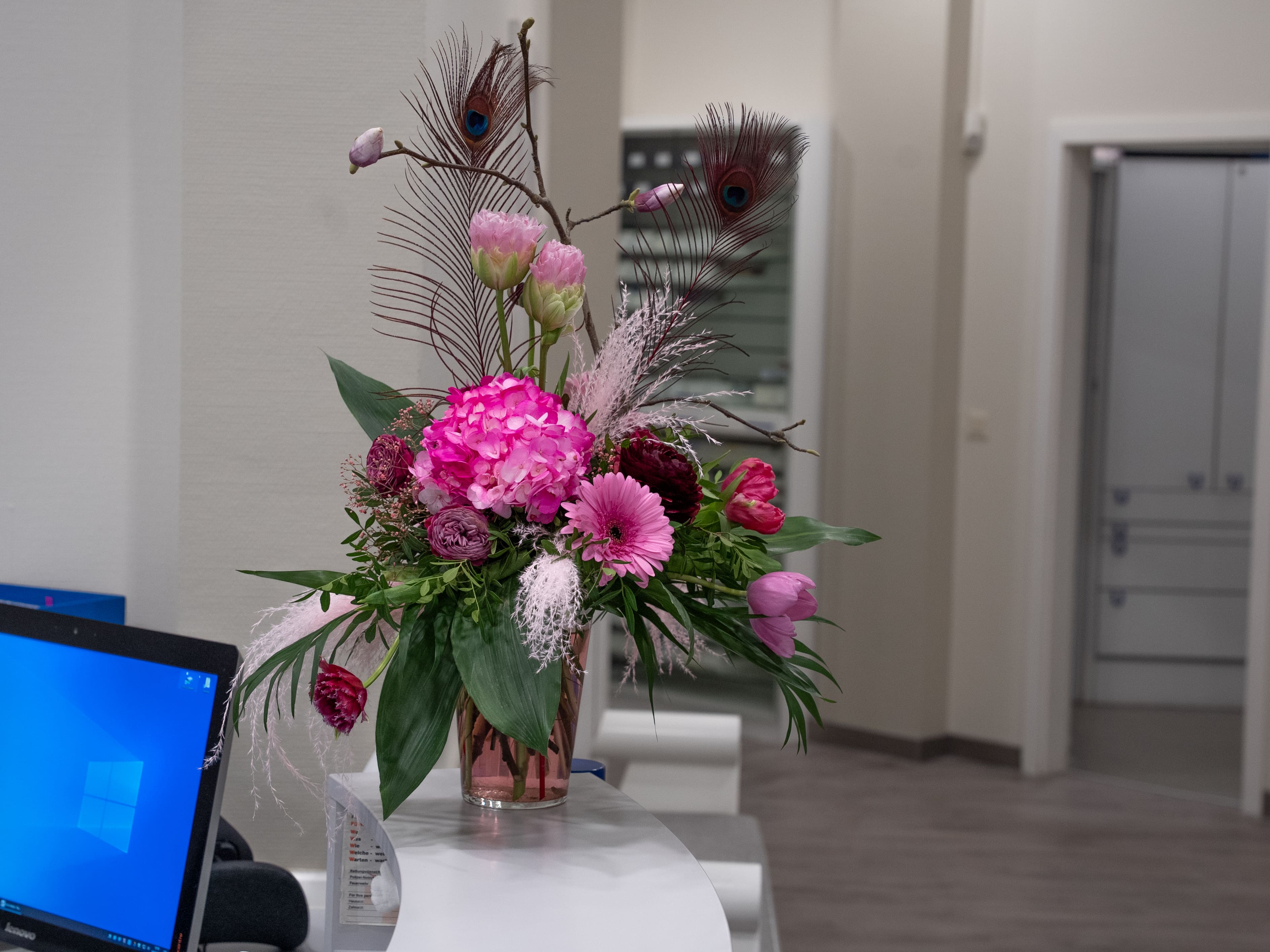 Blumenstrauß mit rosa und lila Blüten, Pfauenfedern, auf hellem Empfangstresen.