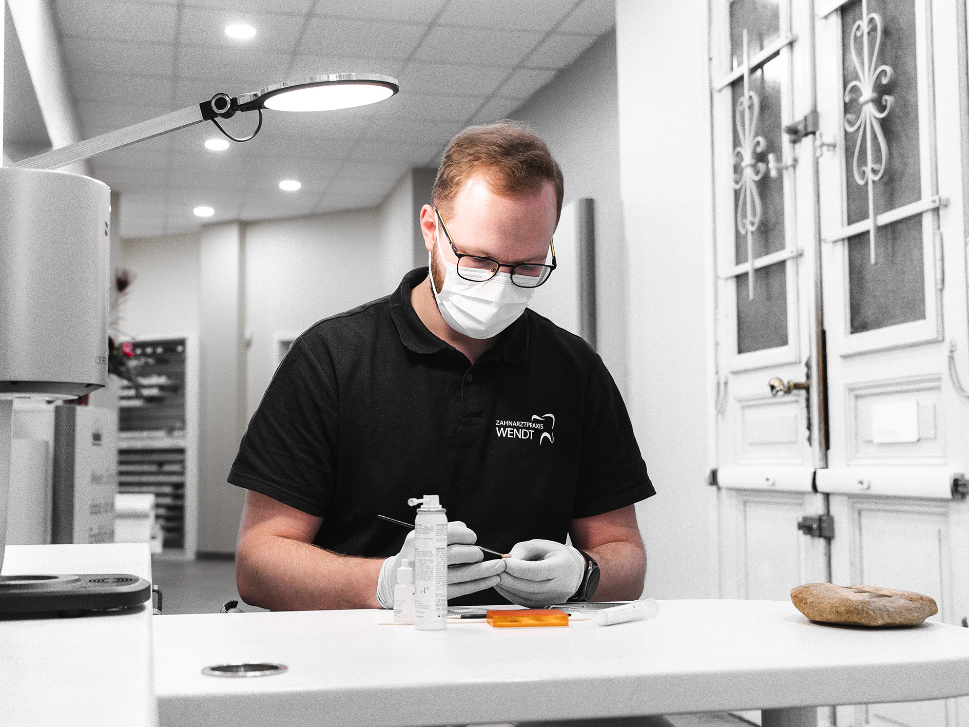 Eine Person mit Maske und Brille arbeitet konzentriert am Tisch in einem modernen Raum.