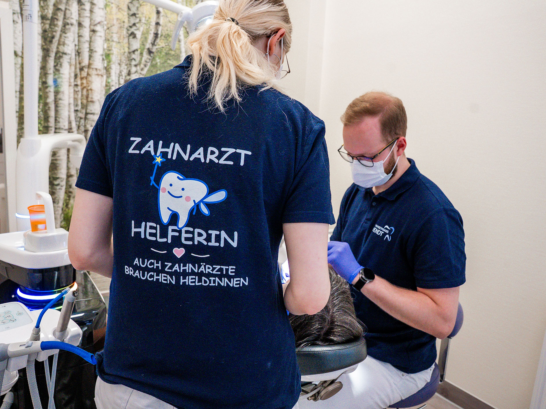 Zahnarzt und Helferin behandeln Patient; beide in blauem Outfit; entspannte Atmosphäre.