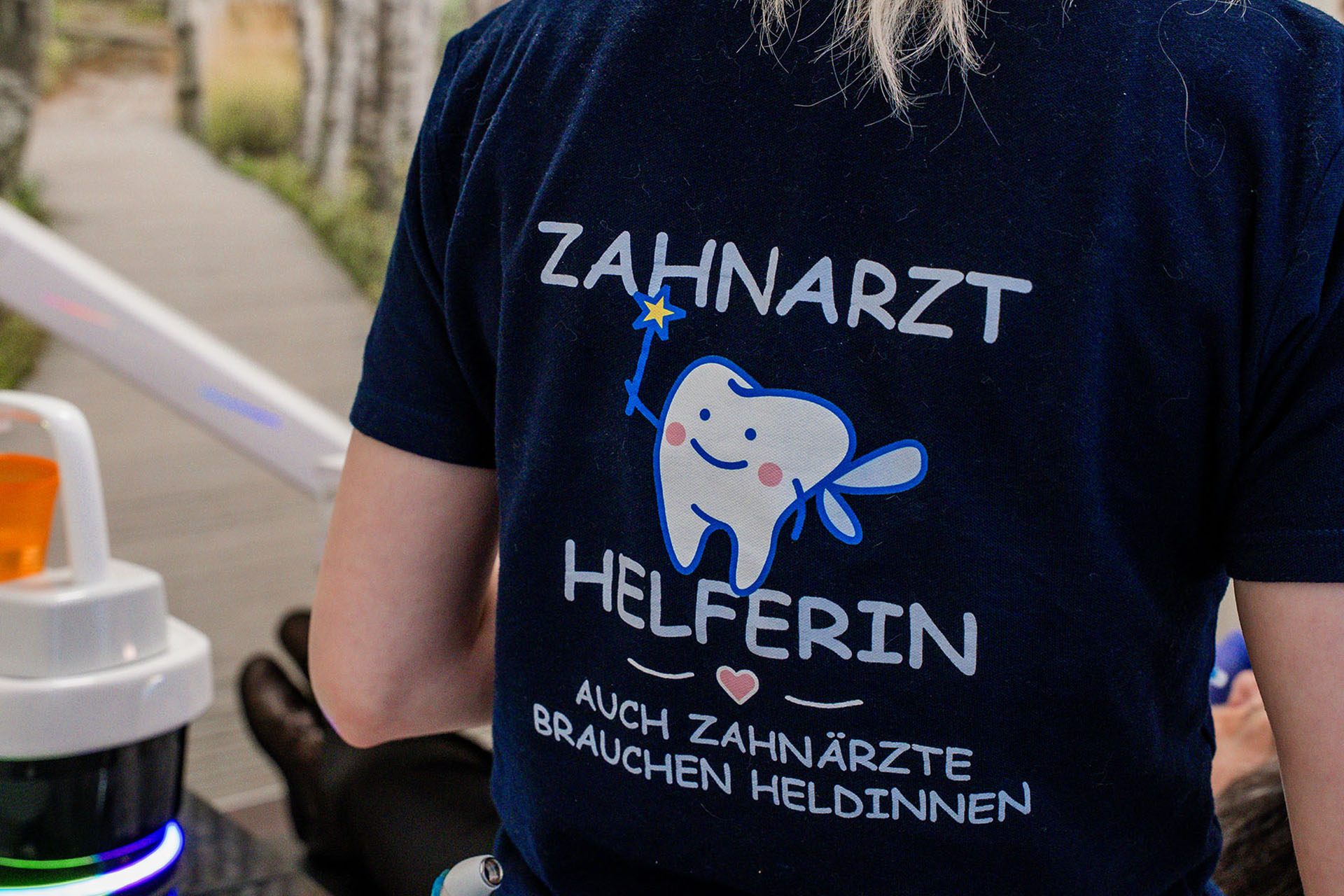 Person in blauem T-Shirt zeigt Cartoon-Zahn mit Zauberstab und der Aufschrift "Zahnarzt Helferin".