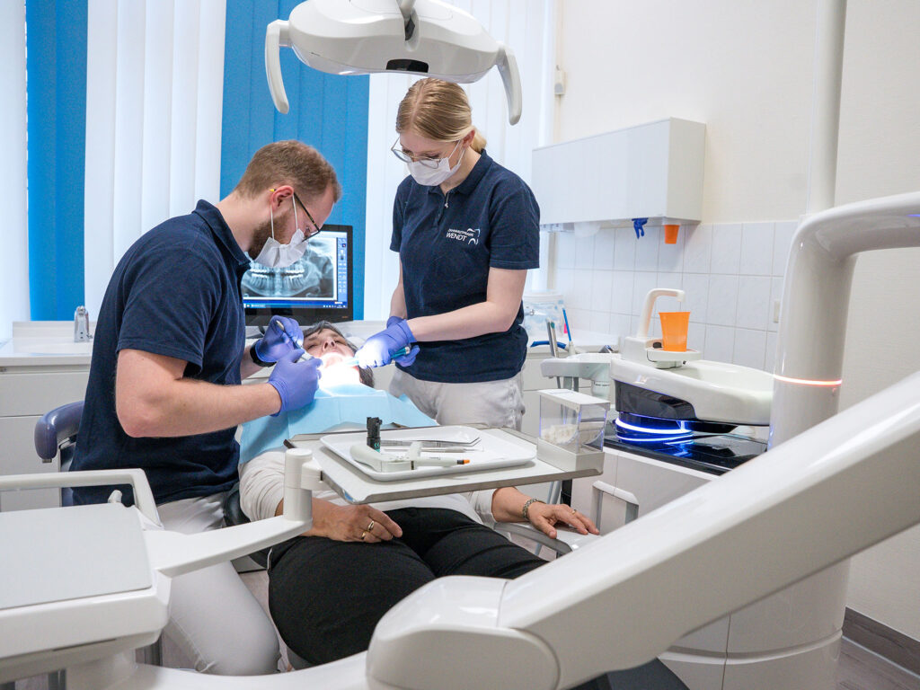 Ein Zahnarzt mit Brille behandelt einen Patienten; eine Assistentin hilft. Modernes Behandlungszimmer.