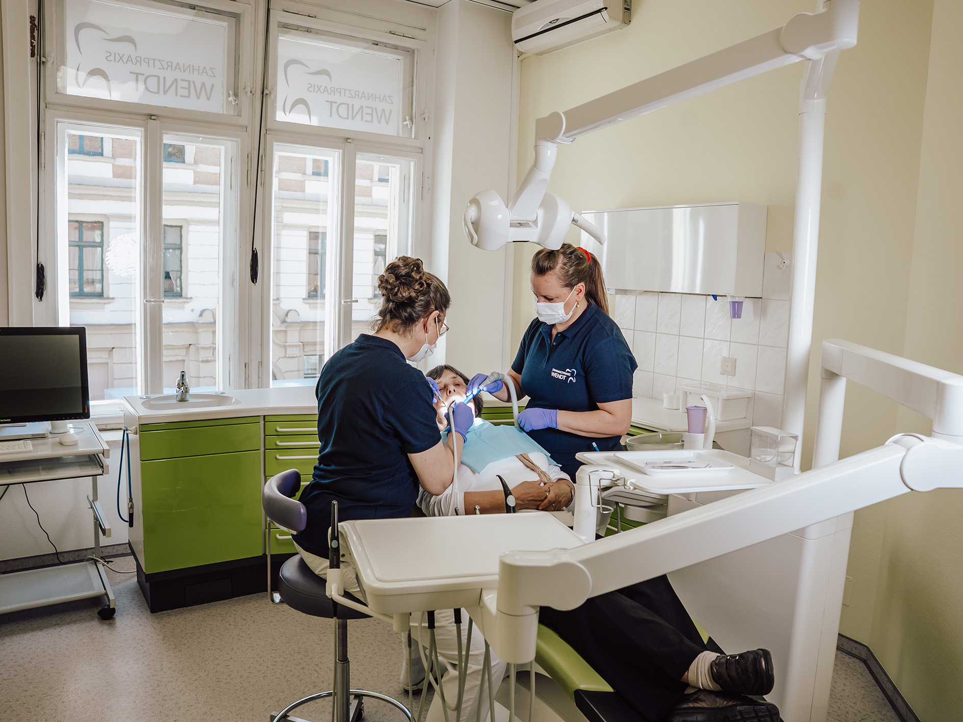Zwei Zahnärztinnen behandeln einen Patienten; helle Praxis, grüne Schränke, Fenster mit Text.