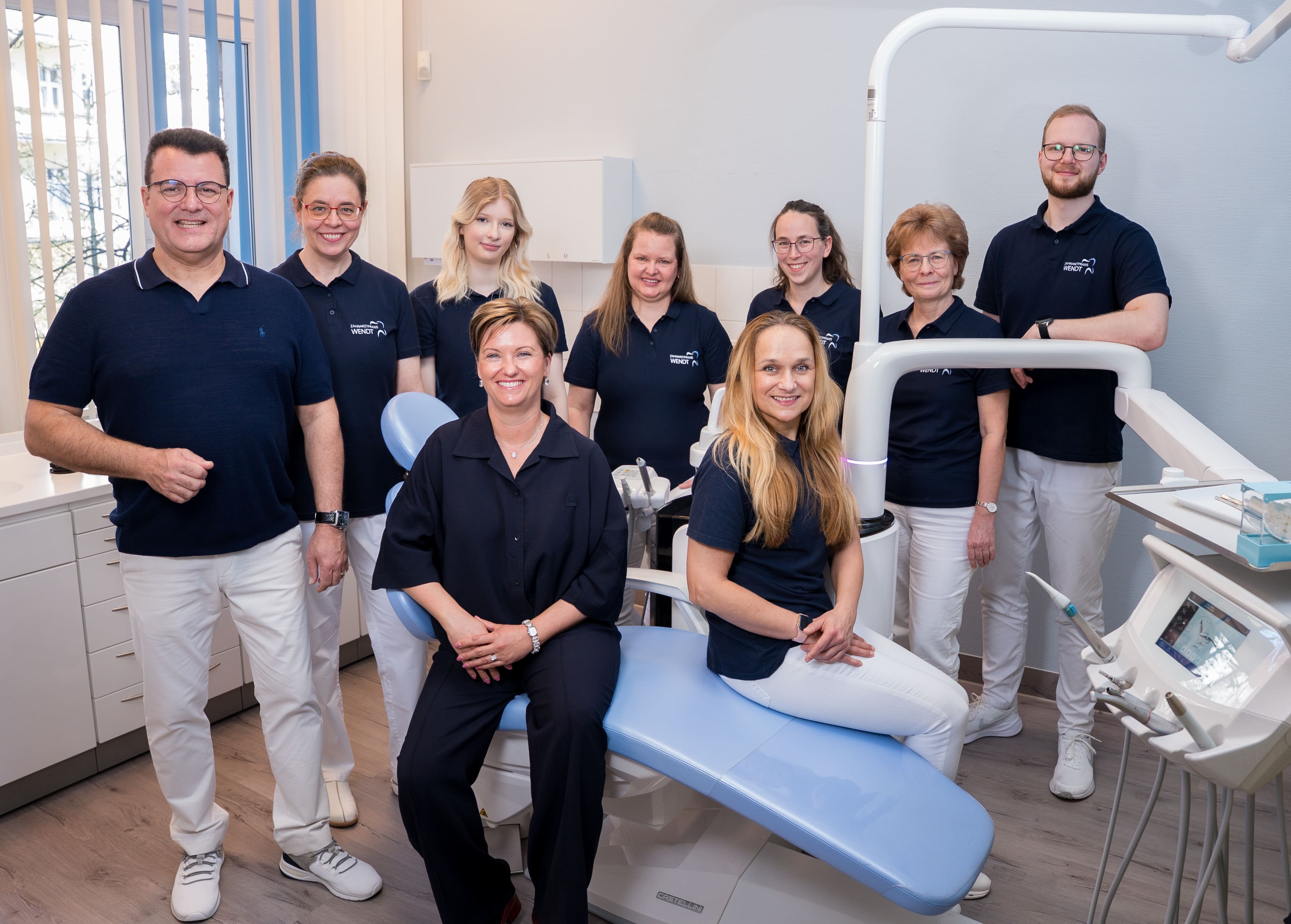 Gruppe von acht Zahnmedizinern in blauer Berufskleidung in einer Zahnarztpraxis. Alle lächeln.