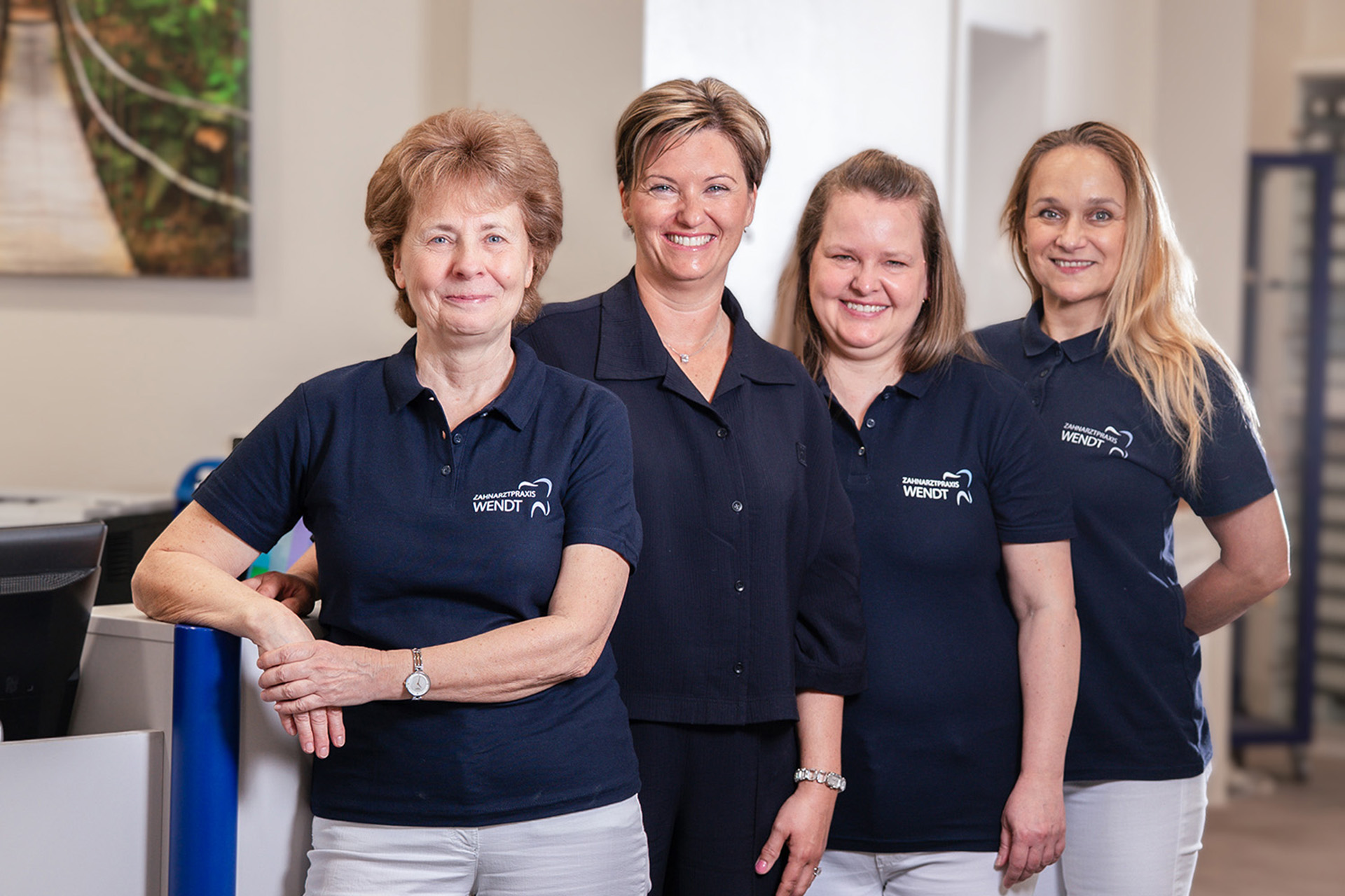 Vier lächelnde Frauen in blauen Poloshirts posieren in einer Büro-Umgebung.
