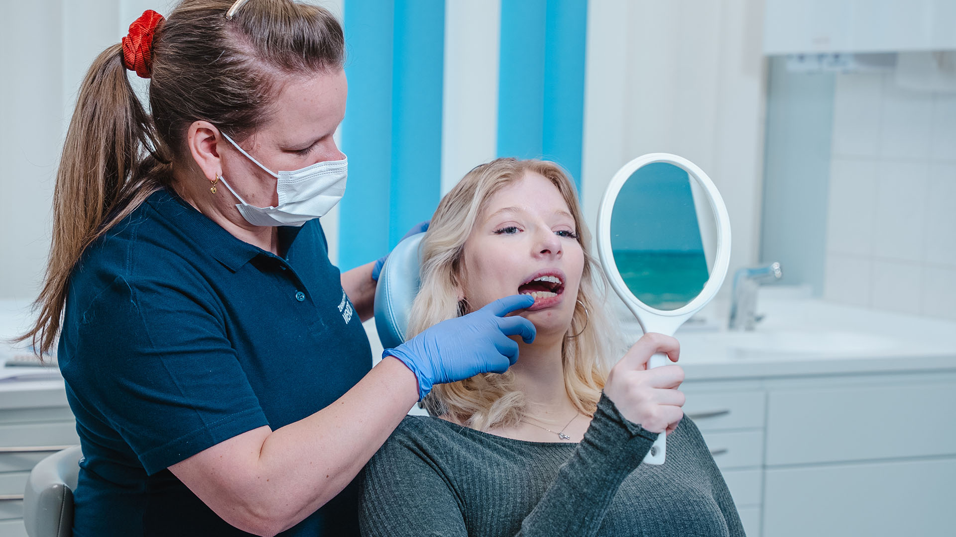 Eine blonde Frau schaut lächelnd mit einem Spiegel auf ihre Zähne beim Zahnarztbesuch