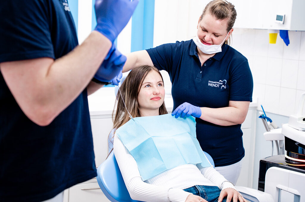 Eine junge Frau mit hellem Haar sitzt lächelnd im Behandlungsstuhl beim Zahnarzt