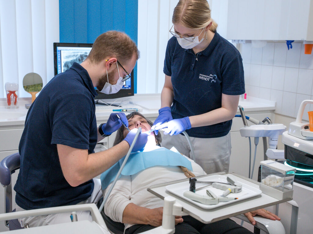 Ein Zahnarzt behandelt einen Patienten und eine Assistentin hilft; helle Praxis, Fenster im Hintergrund.