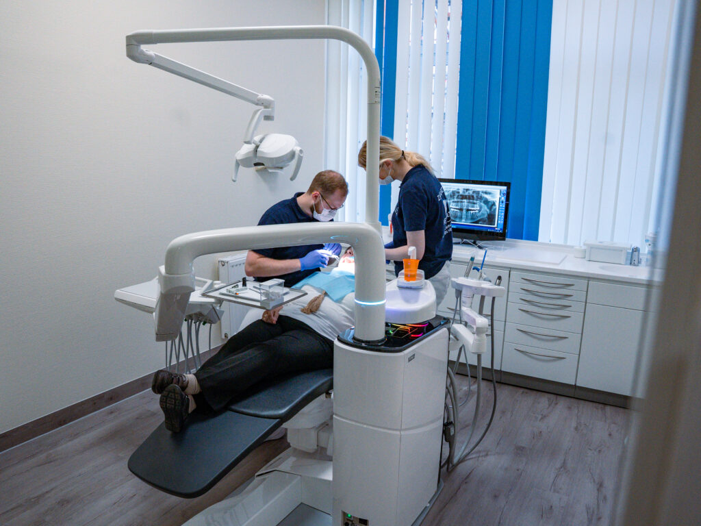 Ein Zahnarzt behandelt einen Patienten und eine Assistentin hilft; helle Praxis, weiße Schränke und Fenster im Hintergrund.